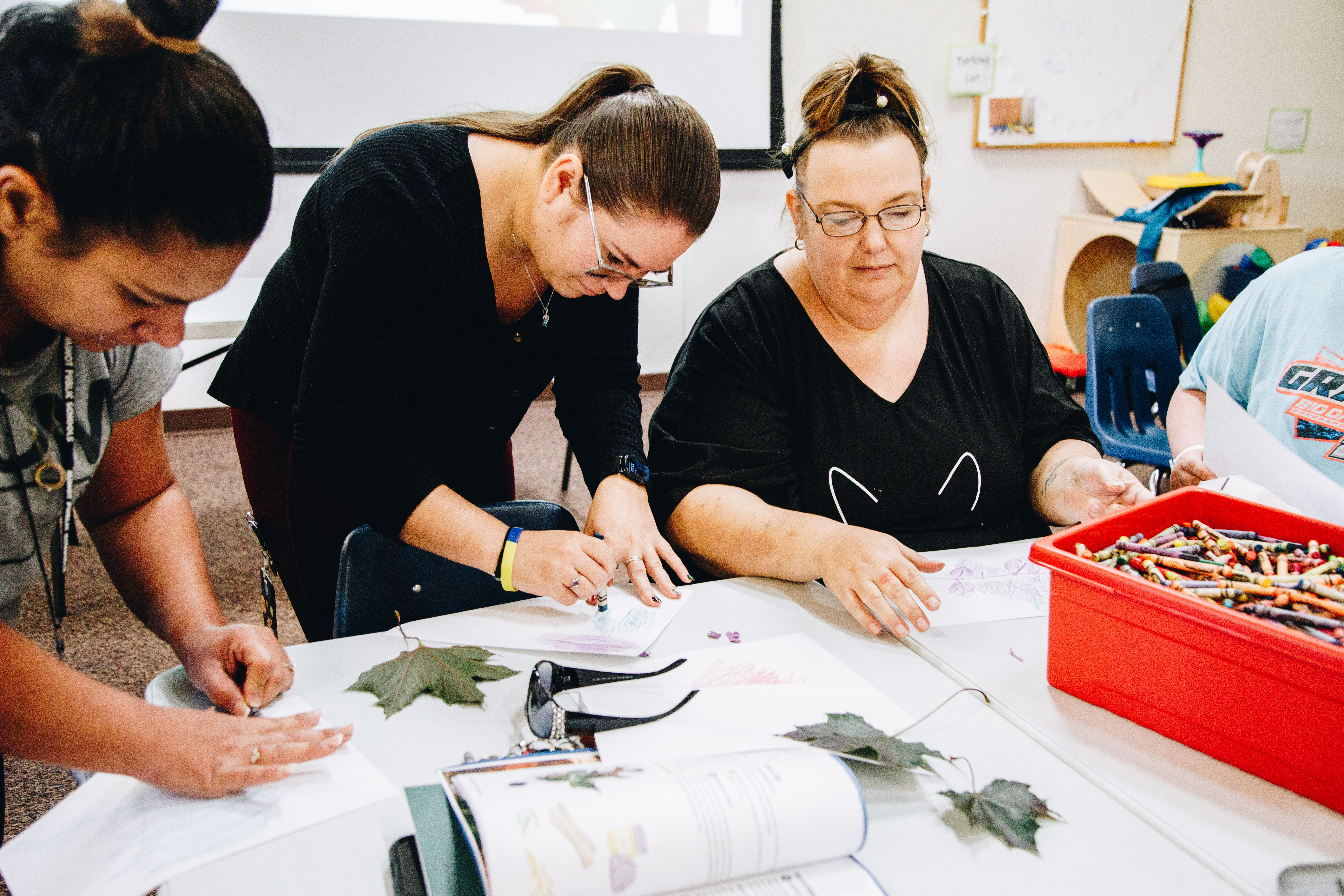 Three women use a crayon to do a leaf rubbing on paper