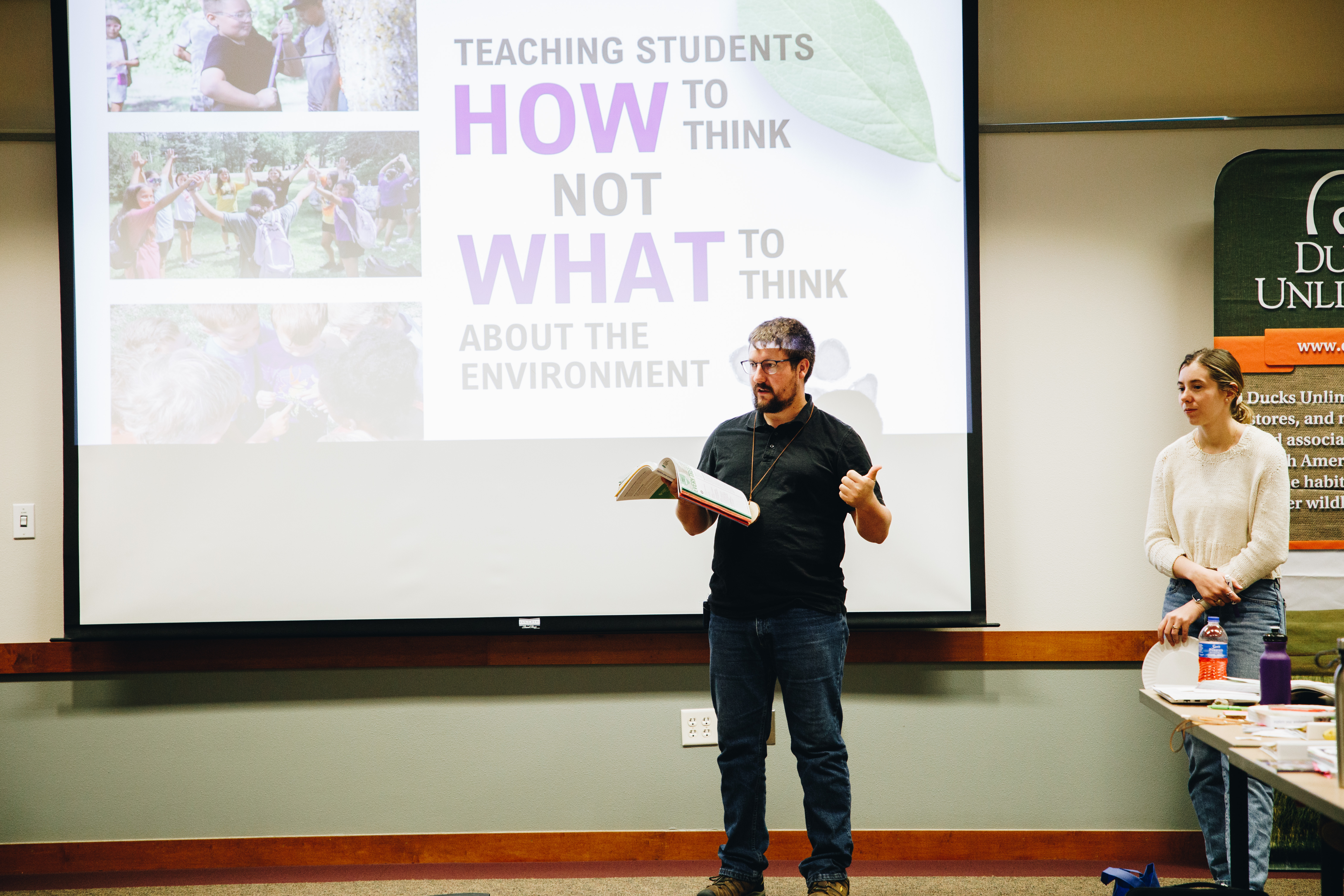 A man reads from a resource book with a project screen behind him that reads "Teaching students HOW to think not WHAT to think about the environment"