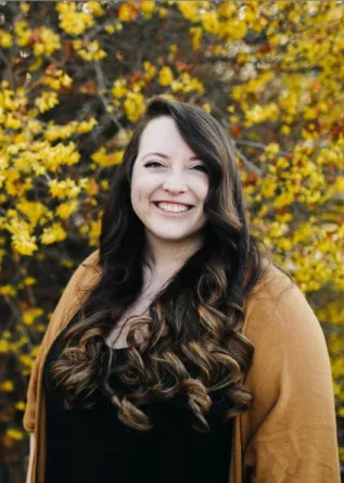 A woman with long, curly brown hair in front of a bush with small yellow flowers