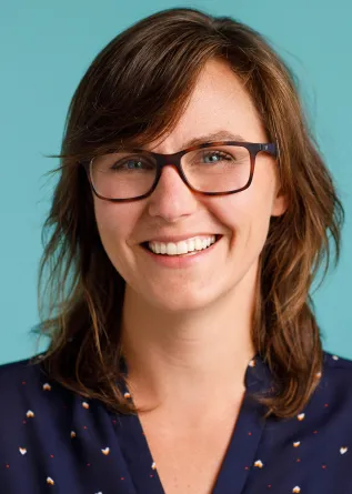 A women standing in front of a vibrant teal background, wearing glasses and a navy blue shirt and glasses.
