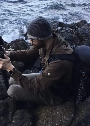 Tyler Waltz sitting on rocks next to a river looking intently at something in his hands.