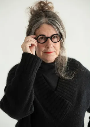 A women in a large black knitted sweater, staring at the camera while touching her large brimmed black circular glasses with her right hand.