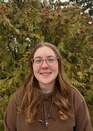 A woman with glasses smiling in front of an evergreen tree