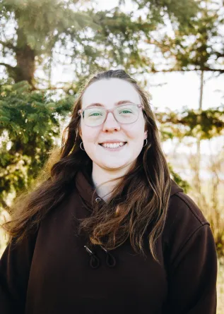 A woman with brown hair and glasses smiles while standing in front of conifer trees