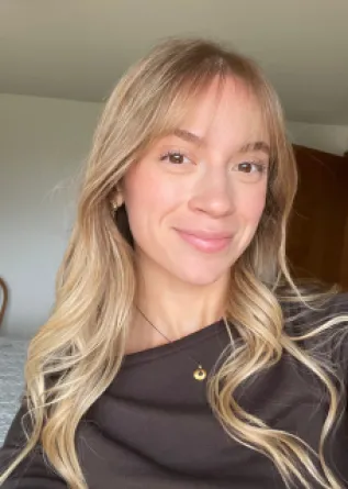 A young woman with long, wavy blonde hair and bangs smiles at the camera, wearing a dark brown off-the-shoulder top.
