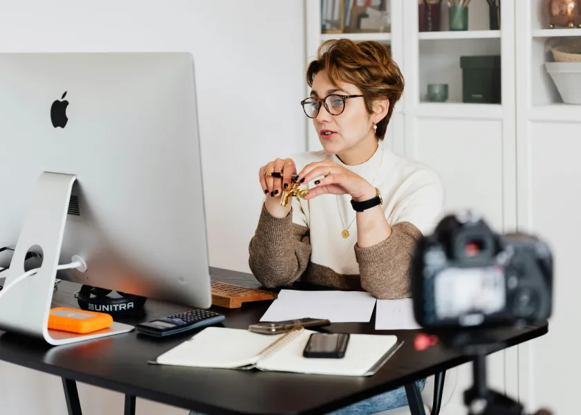 Woman with short hair and glasses sits at desk with computer and a camera filming her in the foreground