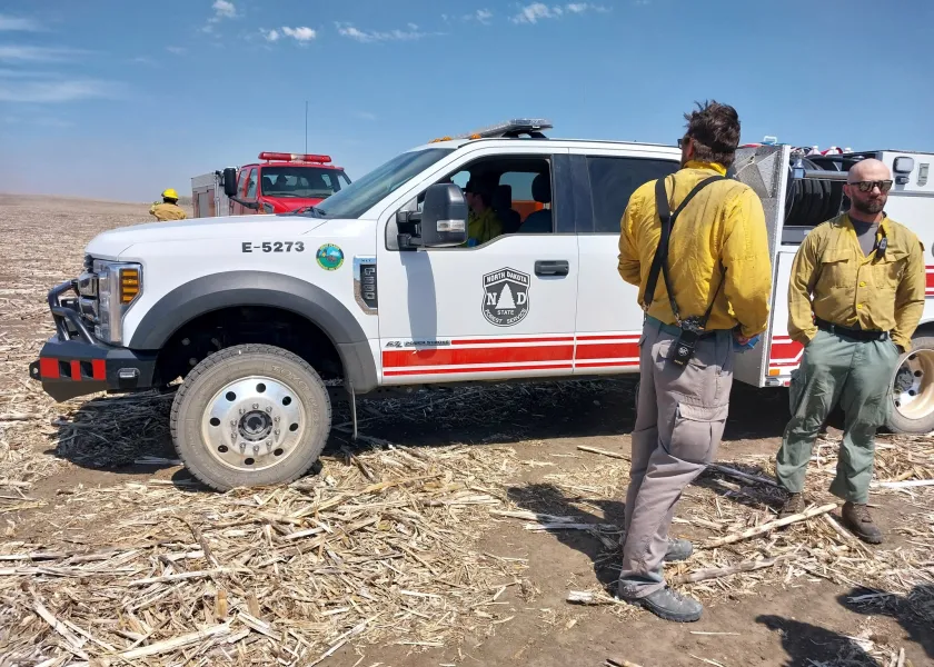 Two firefighters stand in front of a firetruck in a crop field