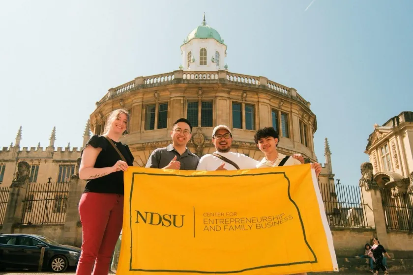 Center for Entrepreneurship and Family Business’s Problems to Possibilities Program Coordinator Jennifer Shillinger, and NDSU students Marcel Roy Domalanta, Mark Rigel Ali and Xyle André Altura stand in Oxford, England while holding a NDSU flag.