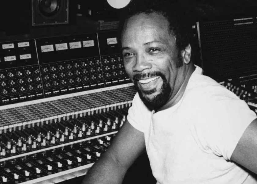Quincy Jones smiling and sitting at a soundboard. 