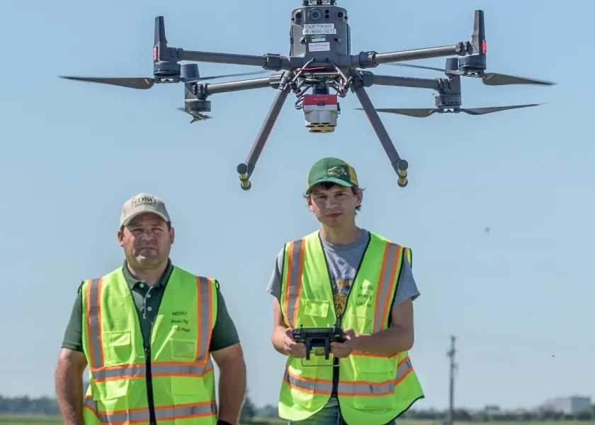 An NDSU professor and student stand in a field with a drone flying above them.