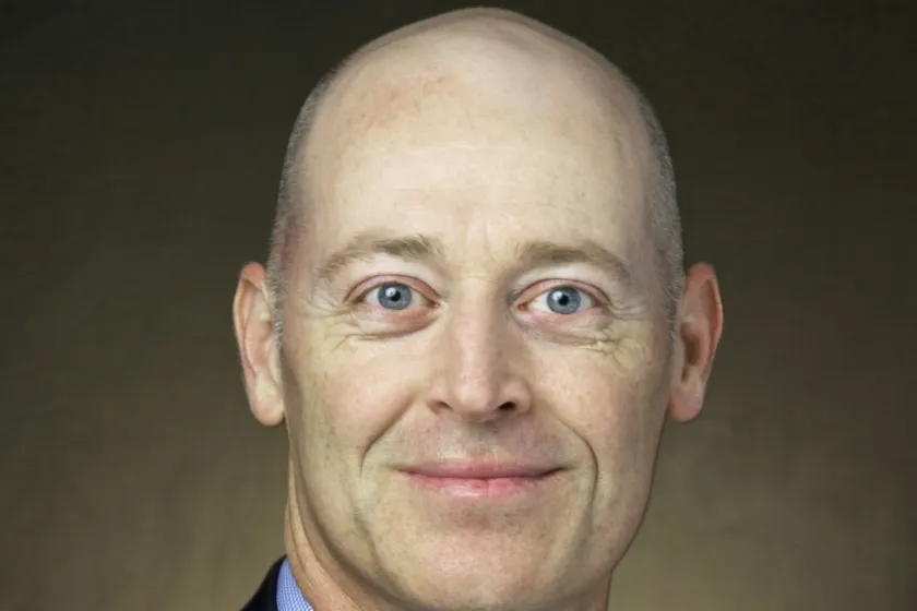 Dr. Tom DeSutter in a blue-collared shirt and suit jacket with a brown background.