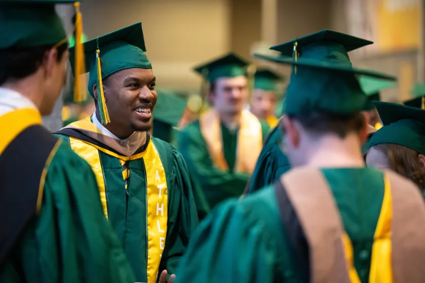 An NDSU student wearing a commencement cap and gown smiles in a crowd of other NDSU graduates.