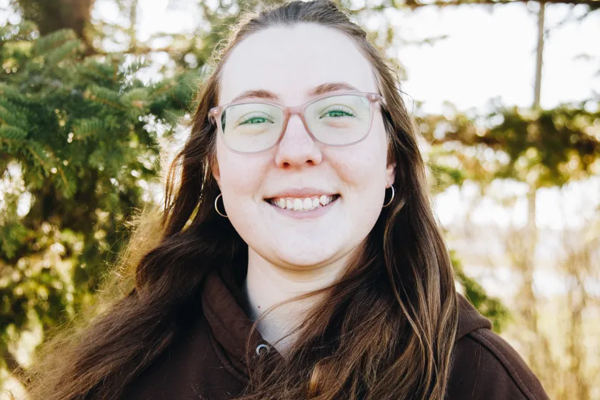 A woman with brown hair and glasses smiles while standing in front of conifer trees