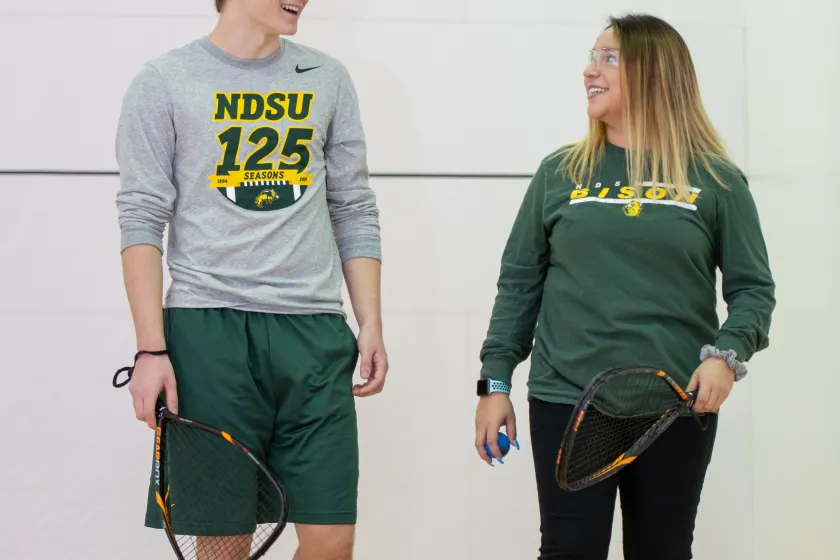 Male student in Bison gear holding racquet talking to woman in Bison shirt and black sweatpants holding racquet