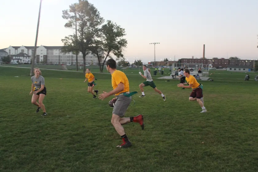 5 students in yellow and grey sporting gear and flag football belts chasing after another student with a football