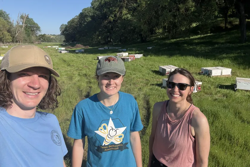 NDSU assistant professor Rogan Tokach standing on grass with two students along with scattered beekeeping equipment in the background.