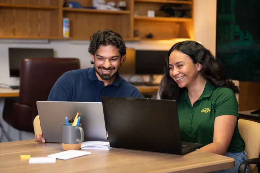 Two students smile as they look at their laptops.