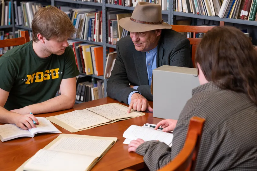 Two NDSU students and a professor sit around a desk to look at a book