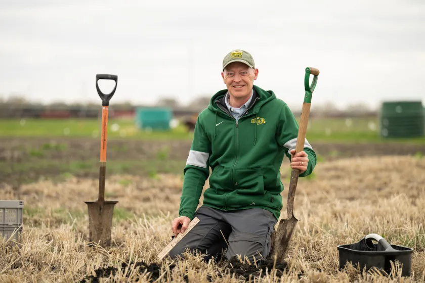 Dr. Tom DeSutter kneeling in a field holding a shovel. 