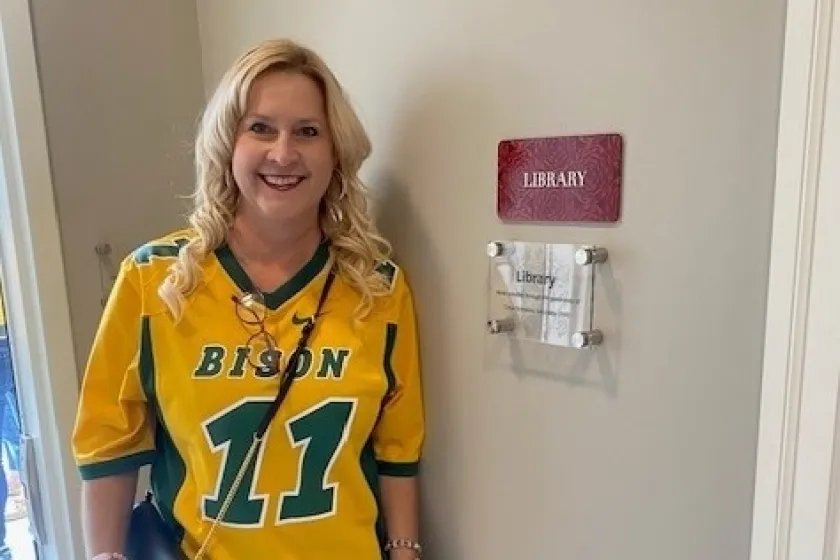 a woman wearing an NDSU jersey stands by a sign that says "library"