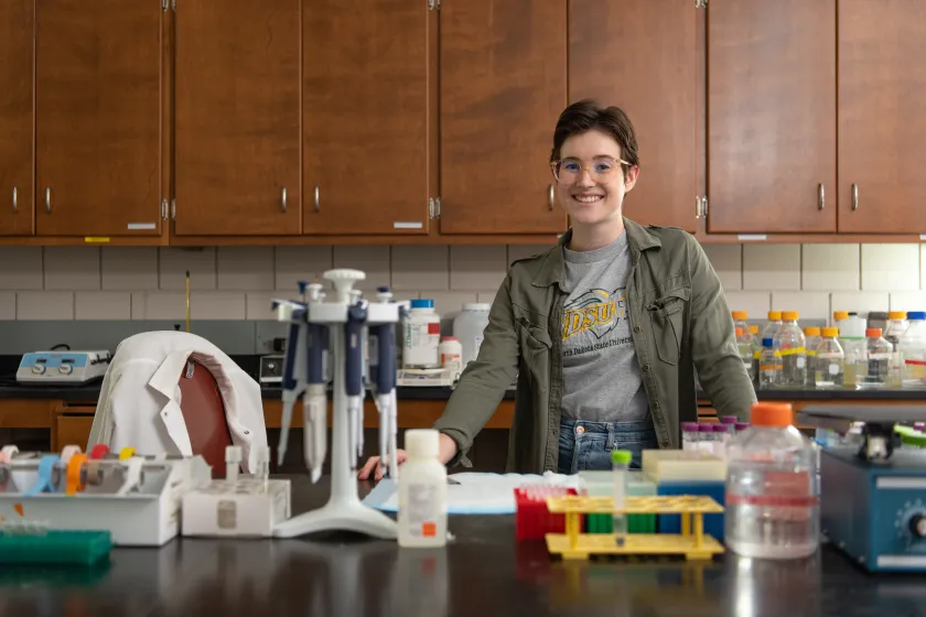 Student smiling confidently into the camera. She is standing in a lab wearing an NDSU shirt and green jacket.