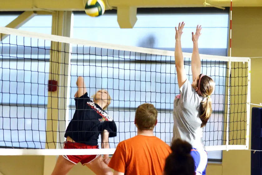 Students playing volleyball. One student is mid-air to spike the ball while another student is mid-air trying to block the spike. Other students are looking on.