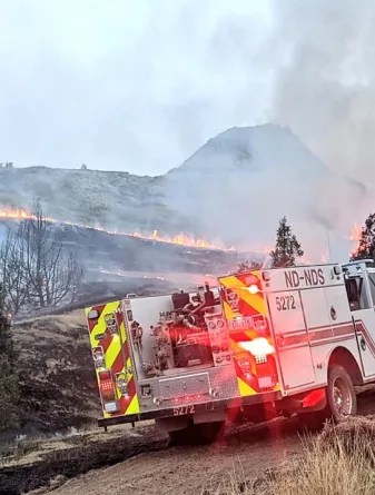 White fire engine with flashing red lights parked near a hill that is on fire with smoke trailing up into the sky