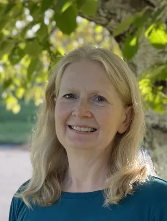 Linda has medium length blonde hair and is wearing a green shirt