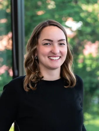 A women with shoulder length brown hair standing in front of a window with trees behind her