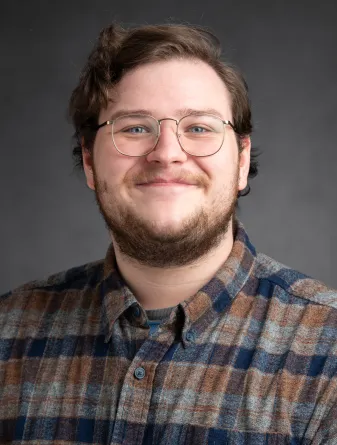 A man with short wavy hair, wearing a buttoned up flannel shirt and a pair of glasses.