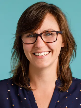A women standing in front of a vibrant teal background, wearing glasses and a navy blue shirt and glasses.