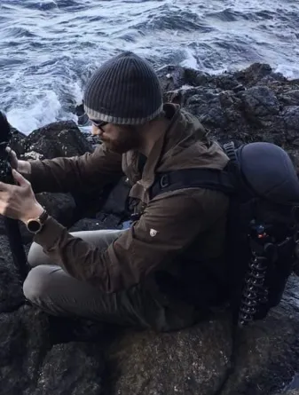 Tyler Waltz sitting on rocks next to a river looking intently at something in his hands.