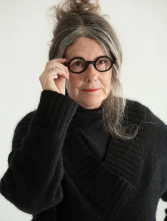 A women in a large black knitted sweater, staring at the camera while touching her large brimmed black circular glasses with her right hand.