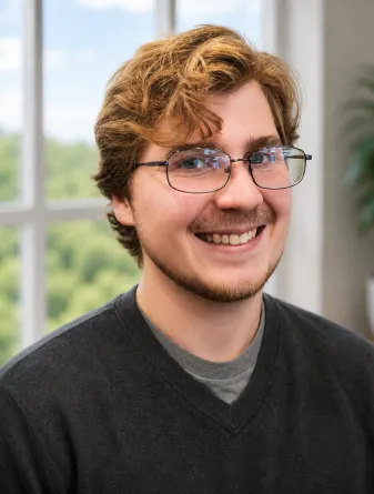 Elijah is a caucasian male with short wavy brown hair with blue eyes and glasses. He is wearing a dark gray shirt. 