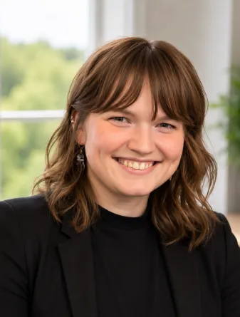 Jordan is a caucasian female young woman with brown shoulder length hair with bangs and brown eyes. She is wearing a black shirt and blazer. 