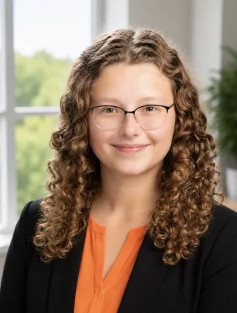 Hania is a caucasian young female with curly long brown hair, brown eyes, and glasses. She is wearing an orange shirt and black blazer. 