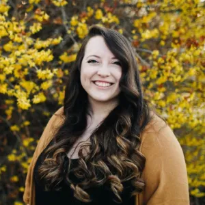 A woman with long, curly brown hair in front of a bush with small yellow flowers