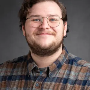 A man with short wavy hair, wearing a buttoned up flannel shirt and a pair of glasses.