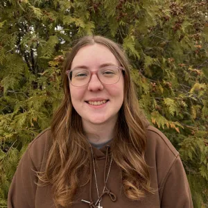 A woman with glasses smiling in front of an evergreen tree