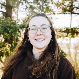 A woman with brown hair and glasses smiles while standing in front of conifer trees