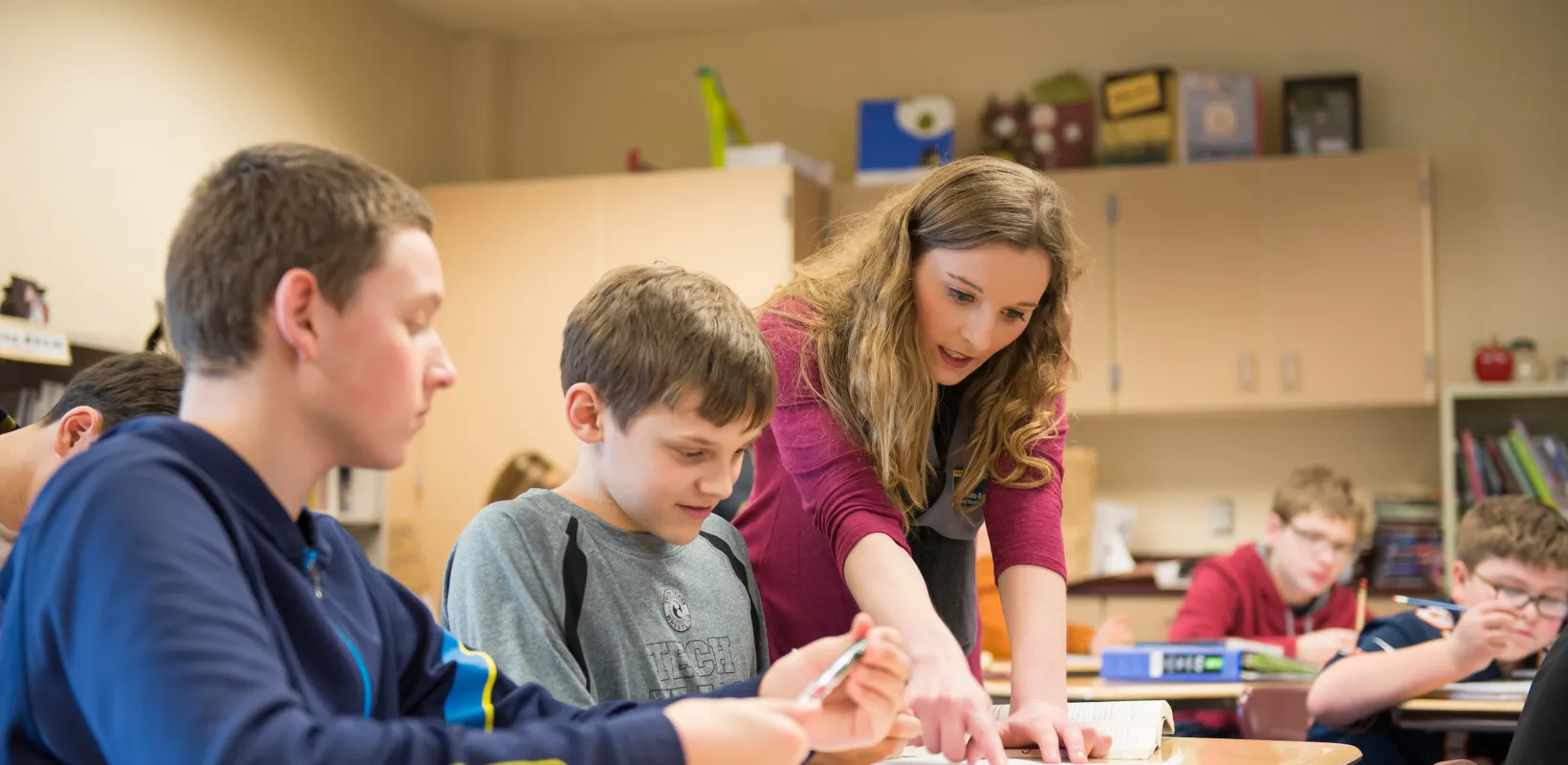 Student teacher leaning on student desk explaining and pointing at students paper.