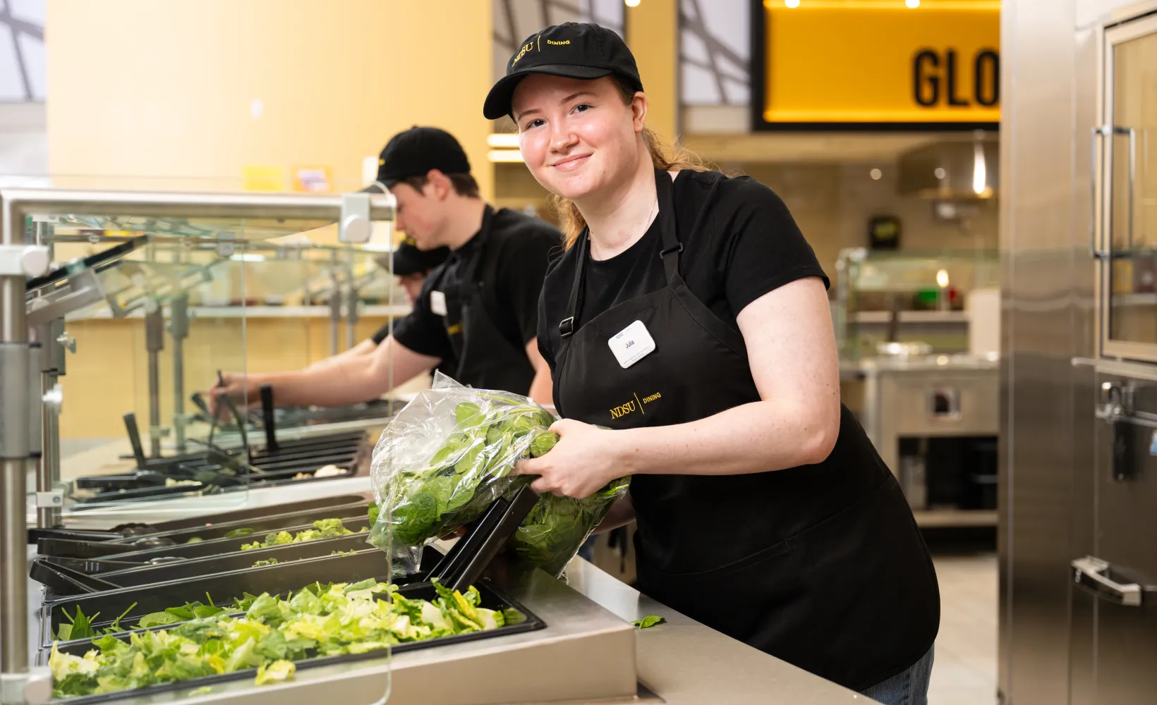 Dining student team members restocking the salad station, while one of the team member pauses from pouring in fresh romaine lettuce to smile for the camera
