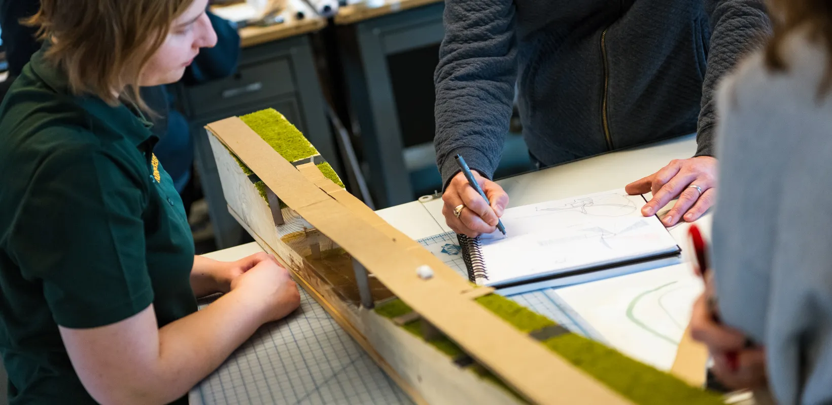Students watching professor as he instructs using pen and paper in design studio.