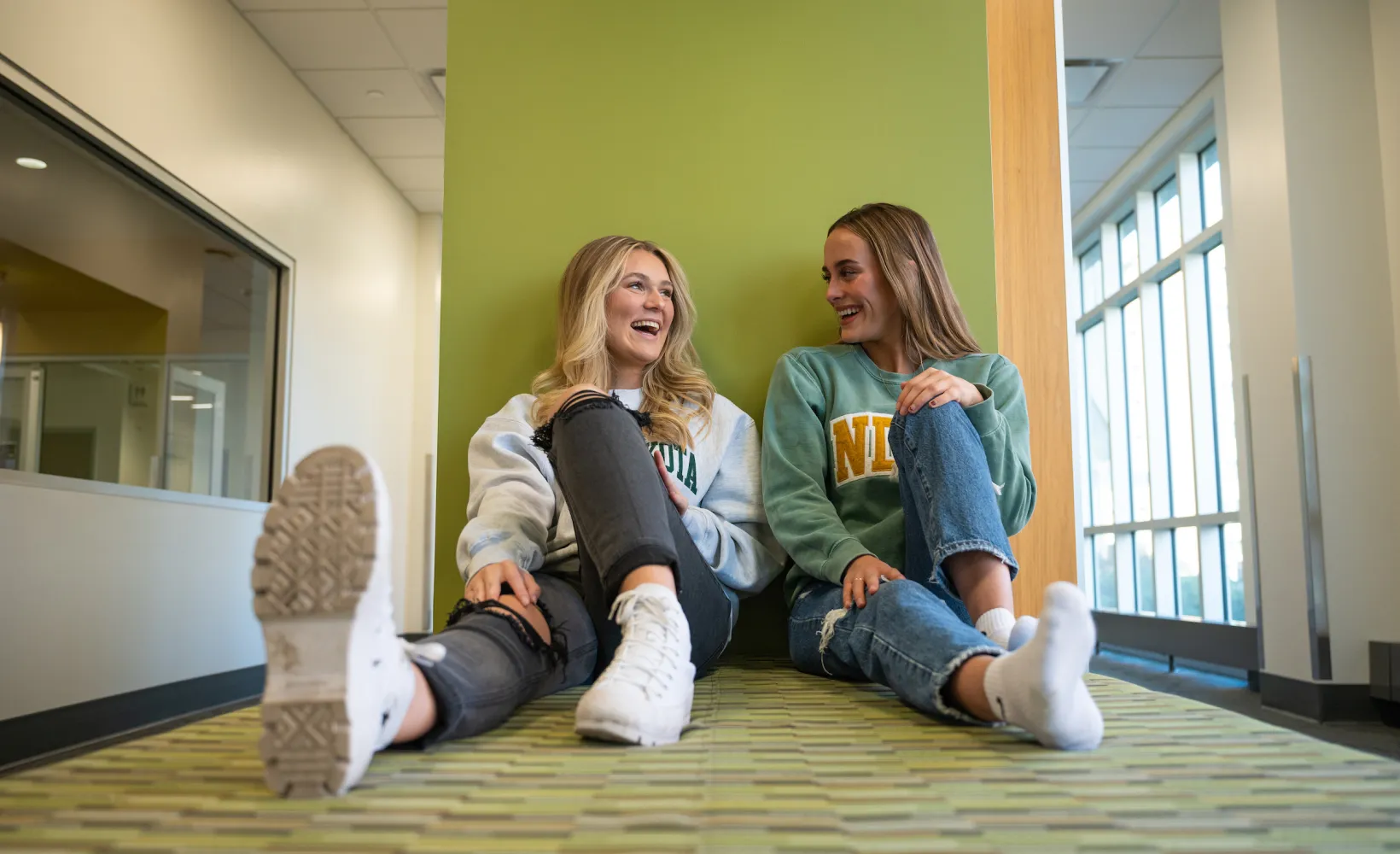 Two students sitting up against a green wall talking and smiling.