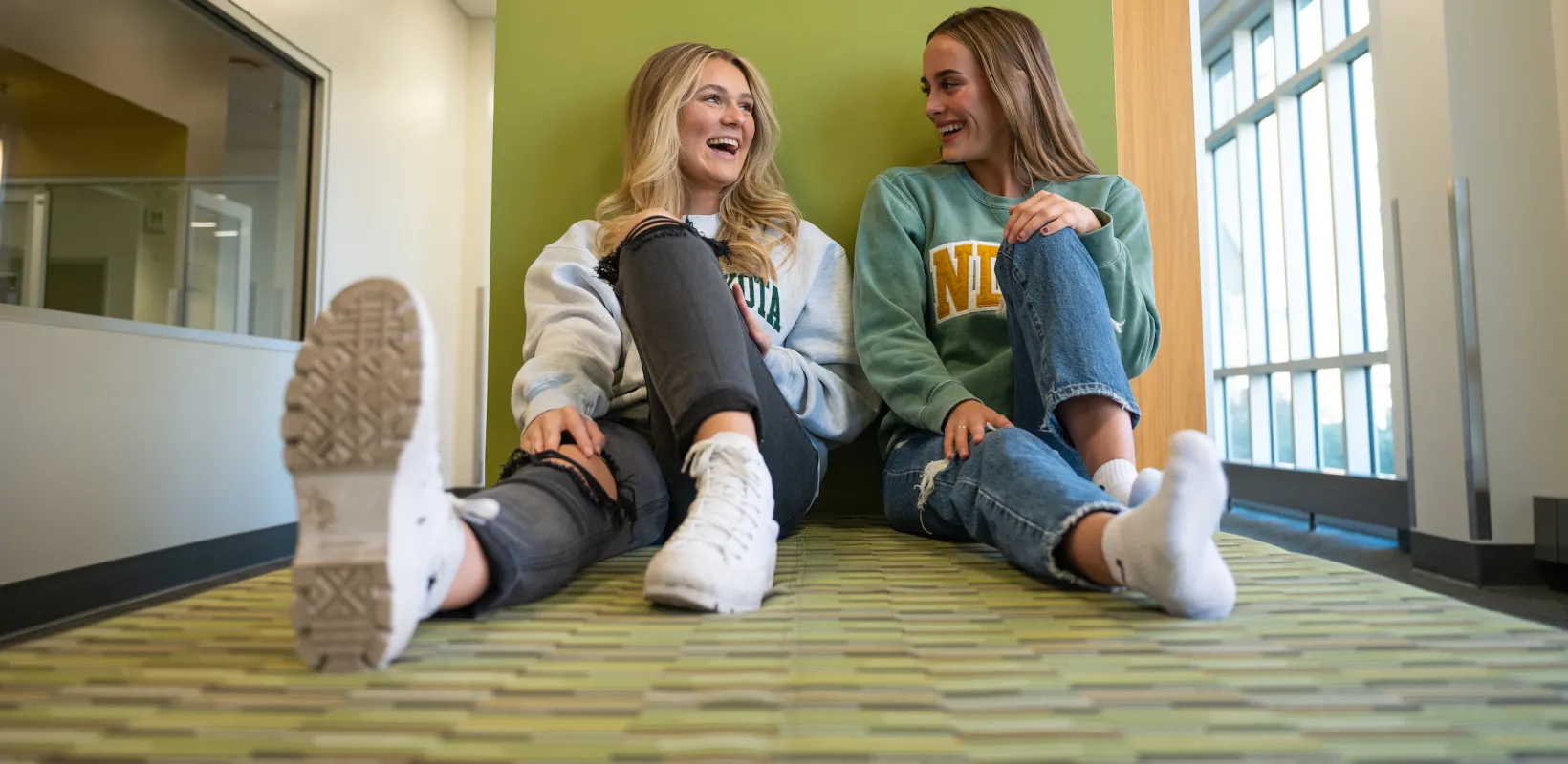 Two students sitting up against a green wall talking and smiling.