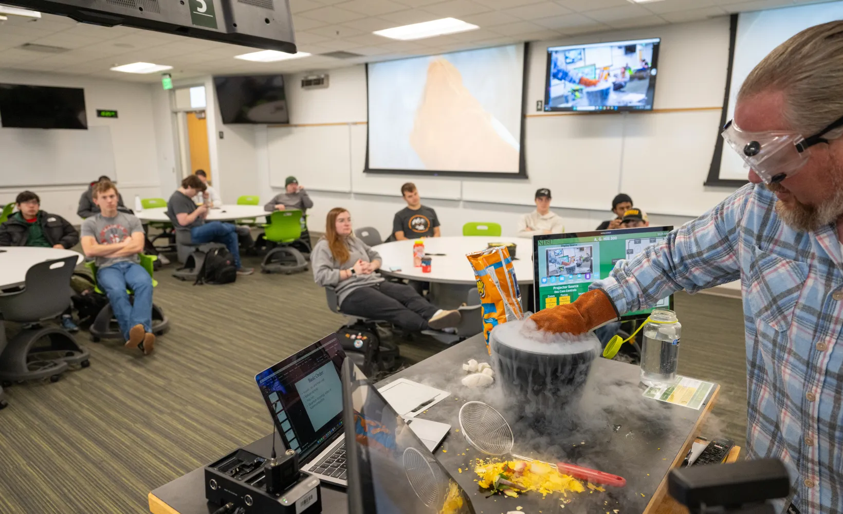 Students watch science experiement conducted by professor at front of classroom.