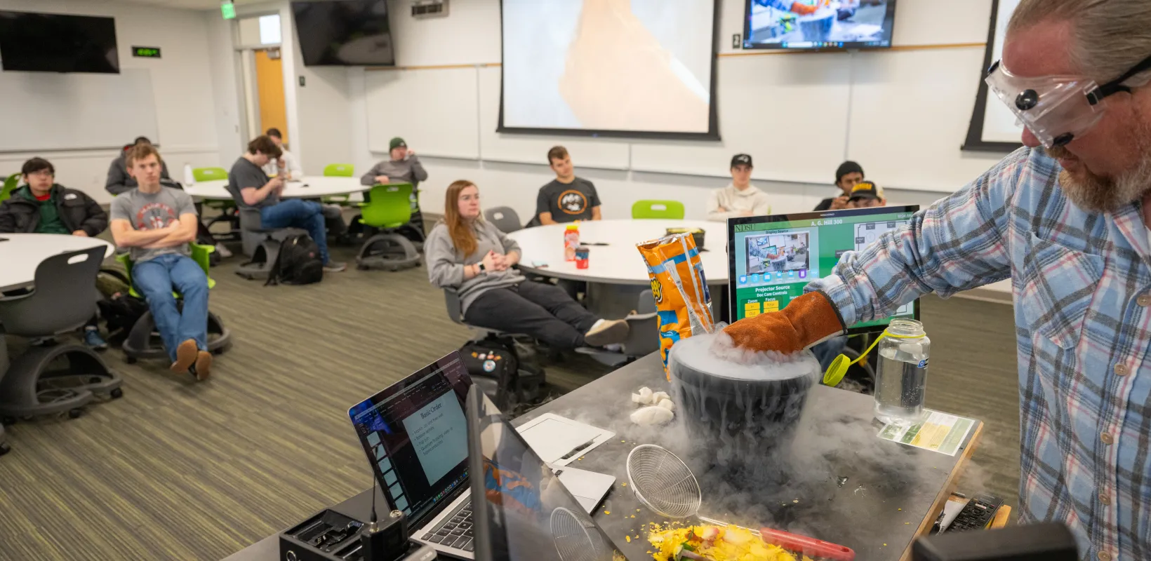 Students watch science experiement conducted by professor at front of classroom.