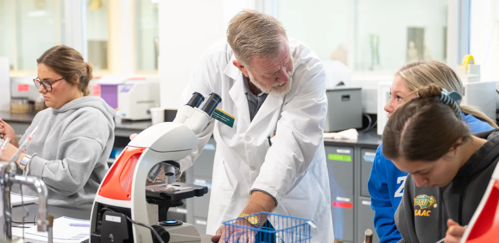 Faculty teaching students in a lab setting with microsopes and other items on lab table.