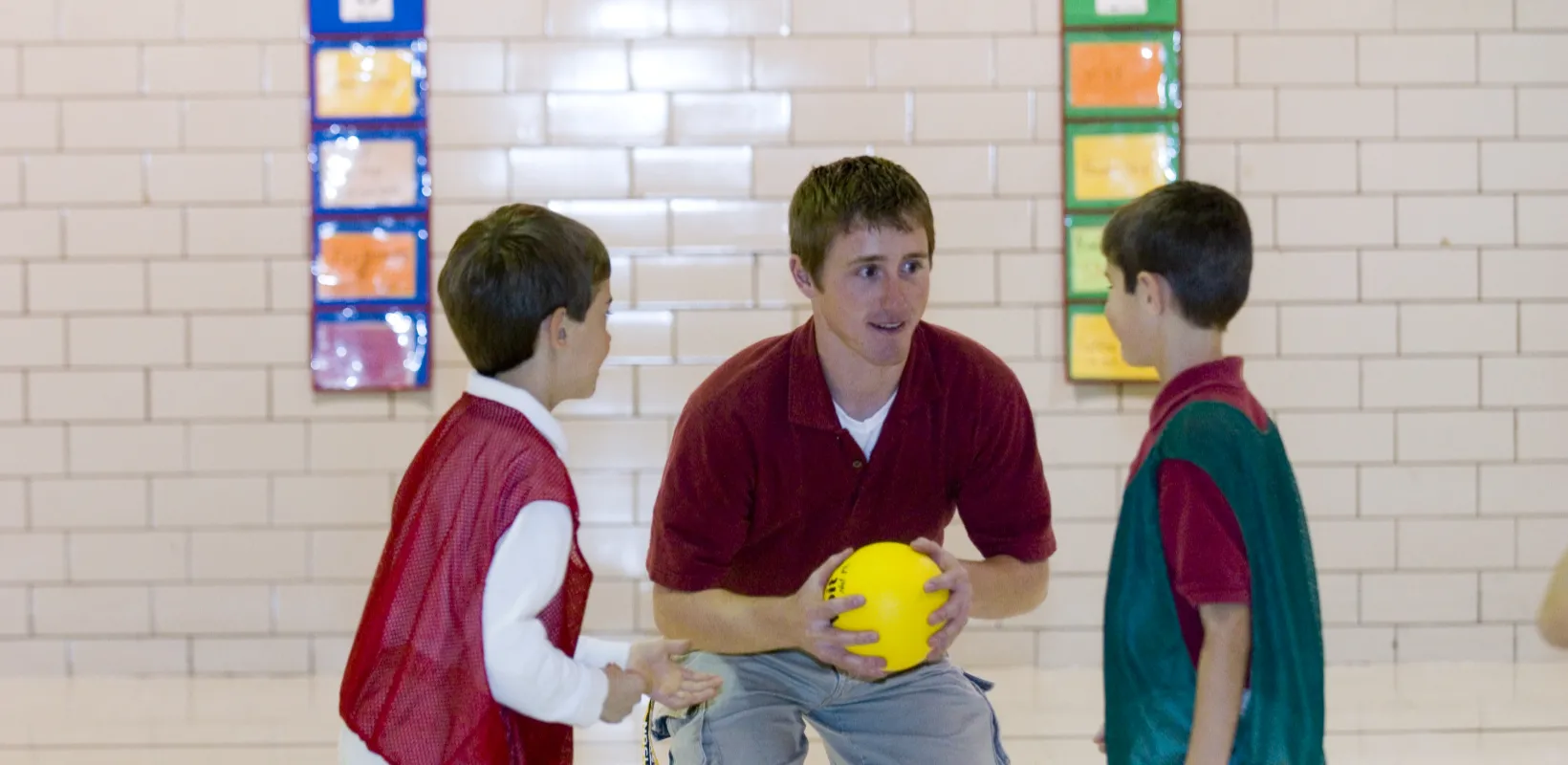 Student teacher working with two students in the gym while holding a yellow ball.