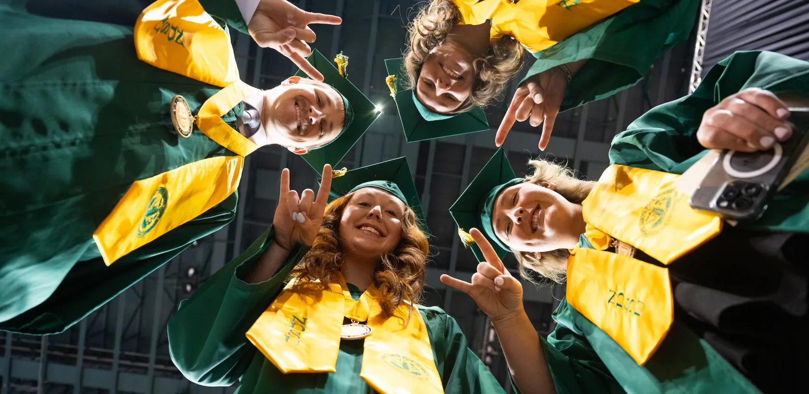 students in a circle at commencement giving bison horns while looking down at photographer